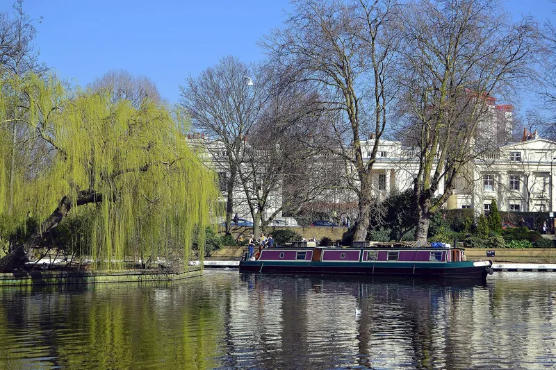 A residential houseboat moored along an urban canal beside trees and historic buildings